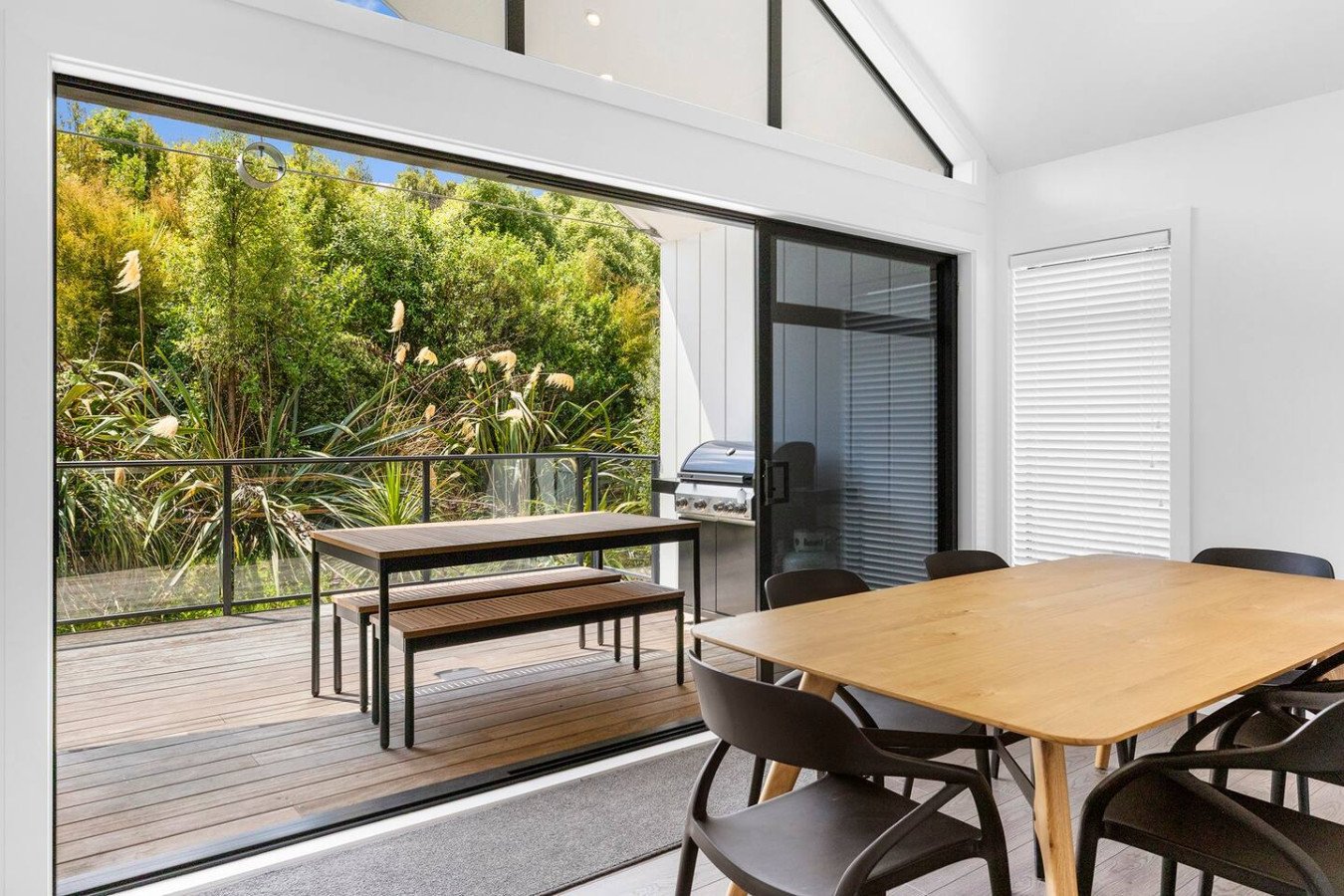 Dining room with a wooden table and black chairs, sliding glass door leading to a deck with a barbecue grill, outdoor table, lush greenery outside, and white walls with blinds.