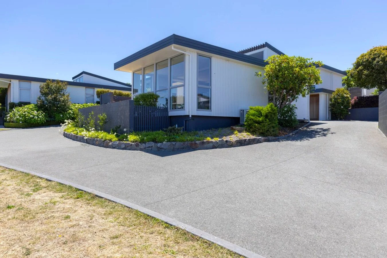 Modern white house with large glass windows, surrounded by greenery, with a grey driveway leading up to it under a clear blue sky.