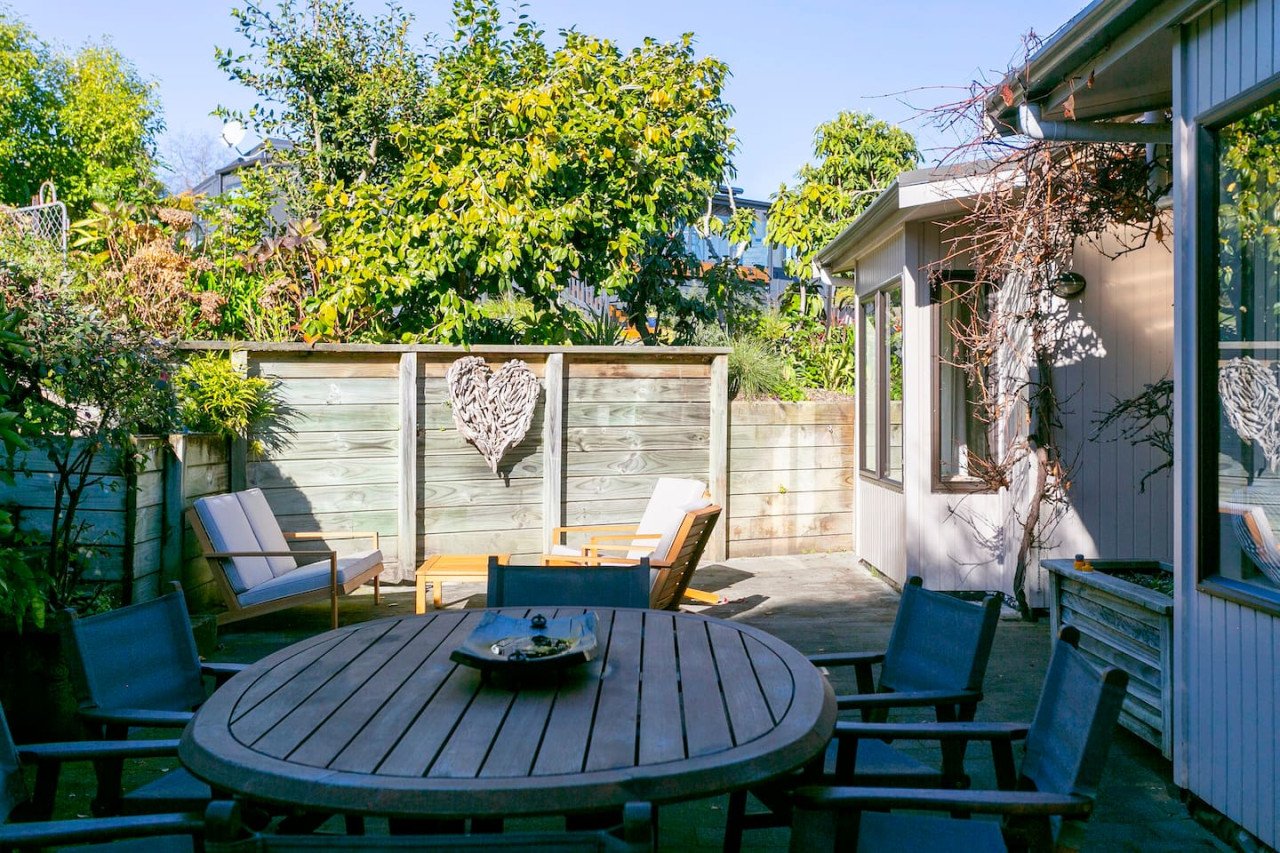A backyard patio with outdoor furniture, including a round table with chairs, a bench, and armchairs. There is a wooden fence with a heart-shaped decoration, trees, and plants in the background, and a small shed with a vine growing on it.
