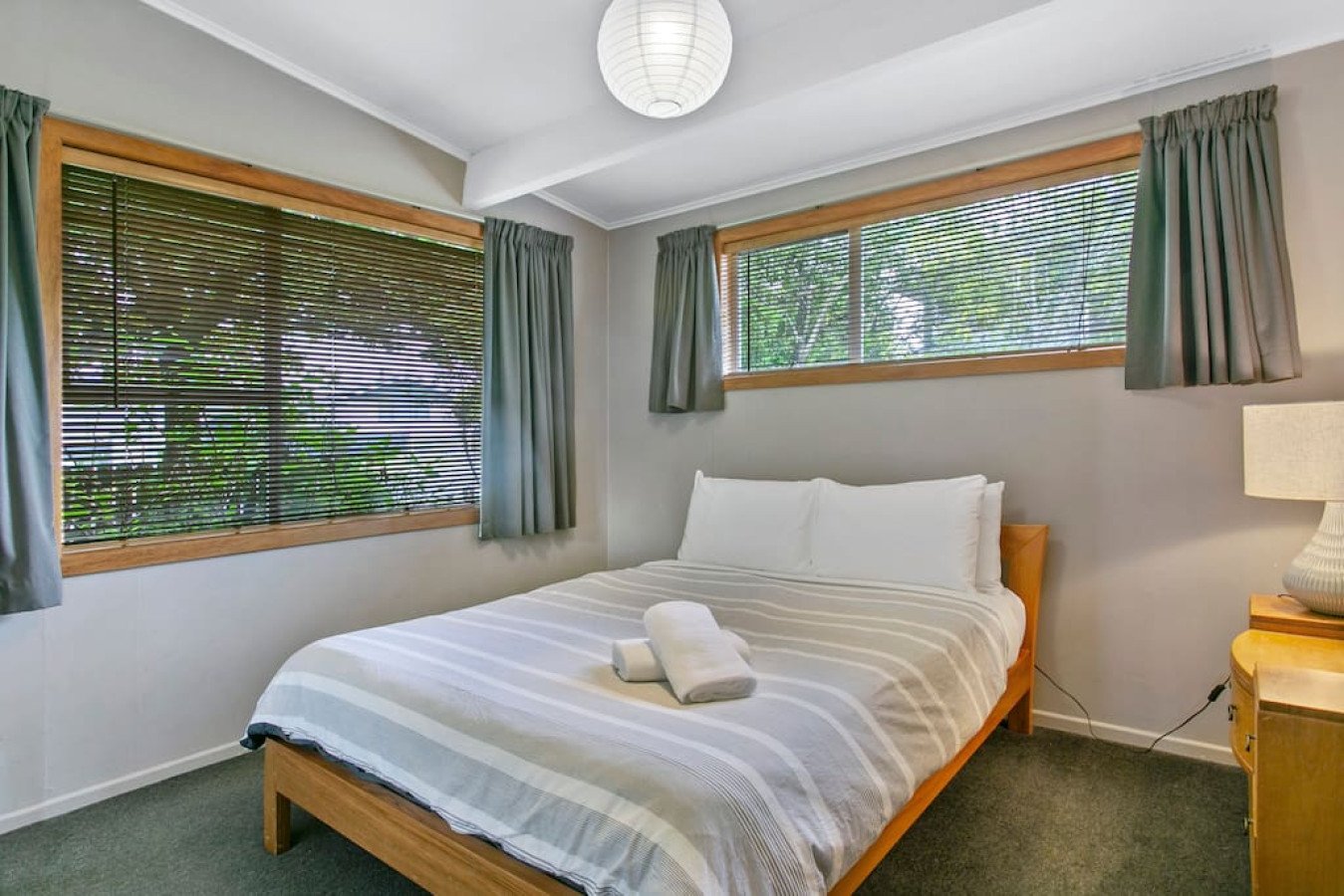 A cozy bedroom with a bed, two rolled towels, green curtains, wooden window frames, and a bedside lamp.