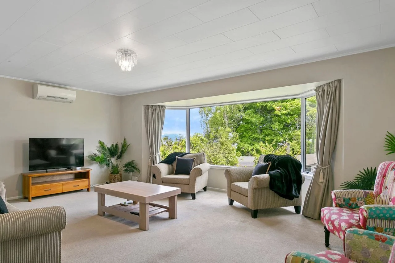Living room with large window, beige sofas, colorful patterned armchairs, a flat-screen TV on a wooden stand, a coffee table, and green houseplants.