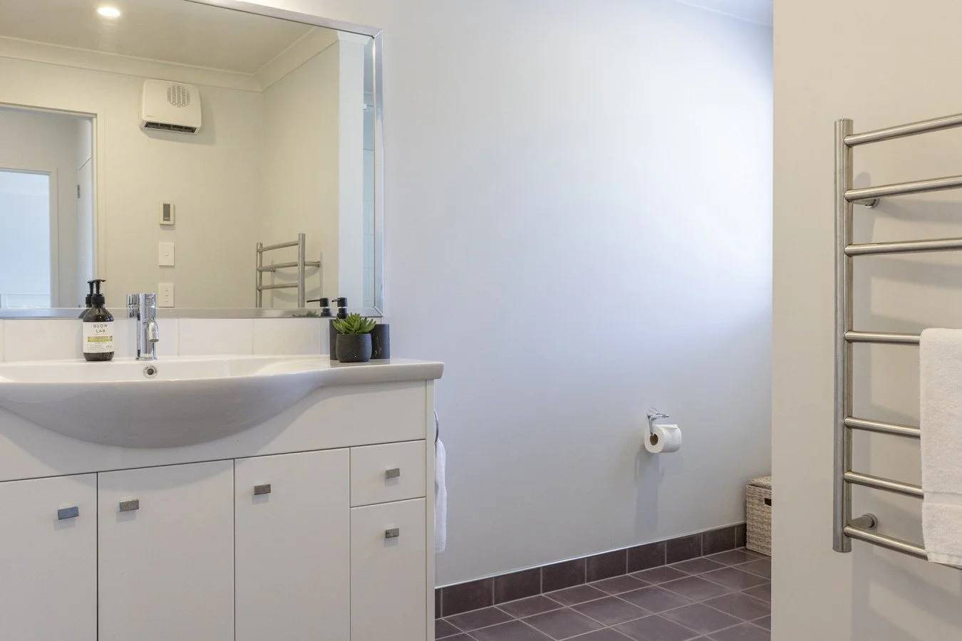 A clean bathroom featuring a white vanity with a sink, a large mirror, a small potted plant, a soap dispenser, a toilet paper roll on the wall, a towel rack with a towel, and a basket in the corner. The bathroom has white walls, dark tile flooring, a