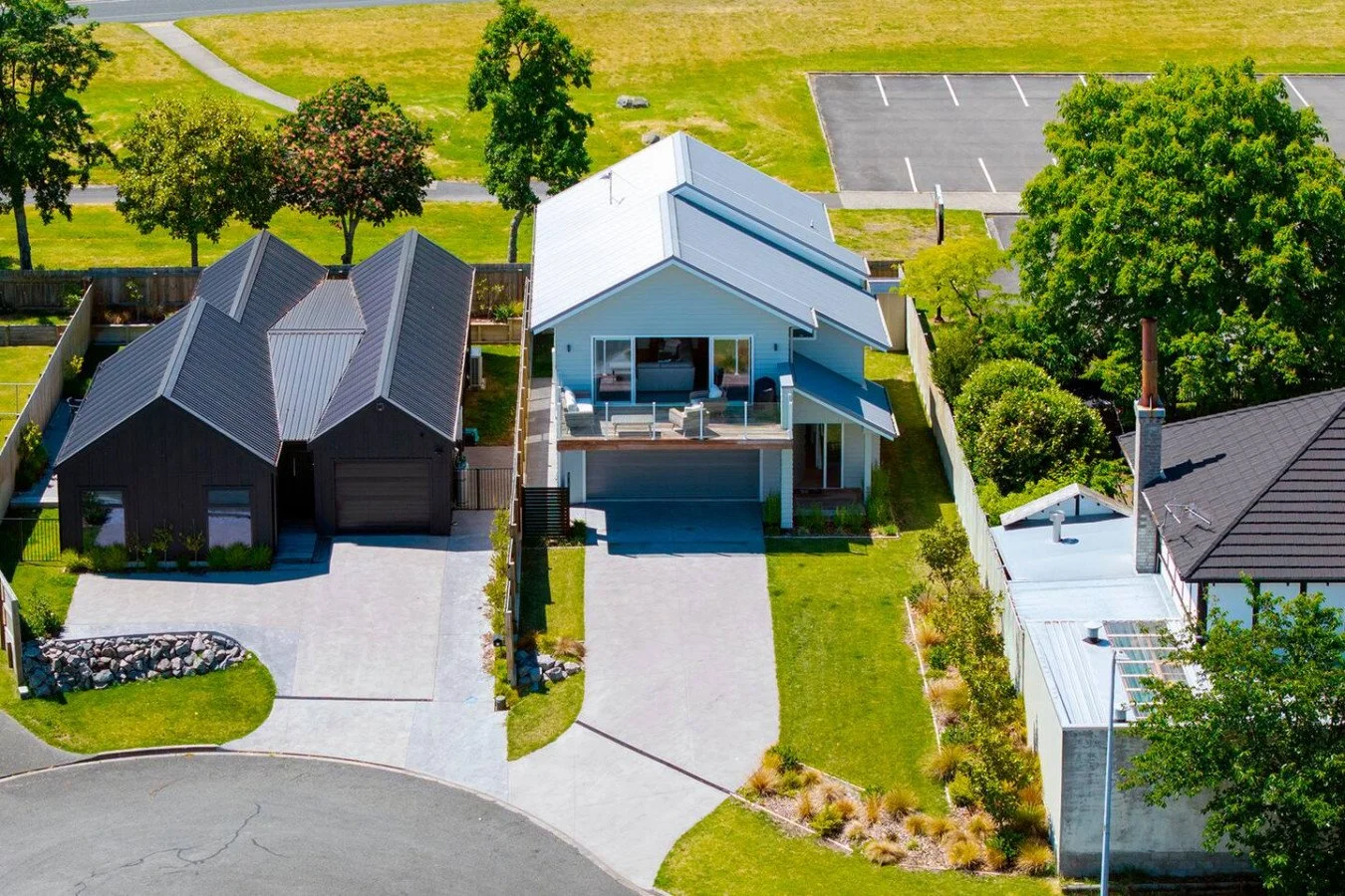Aerial view of a modern house with a white exterior, large balcony, and green lawn, neighboring a black house with a garage, on a quiet street with trees and park in the background.