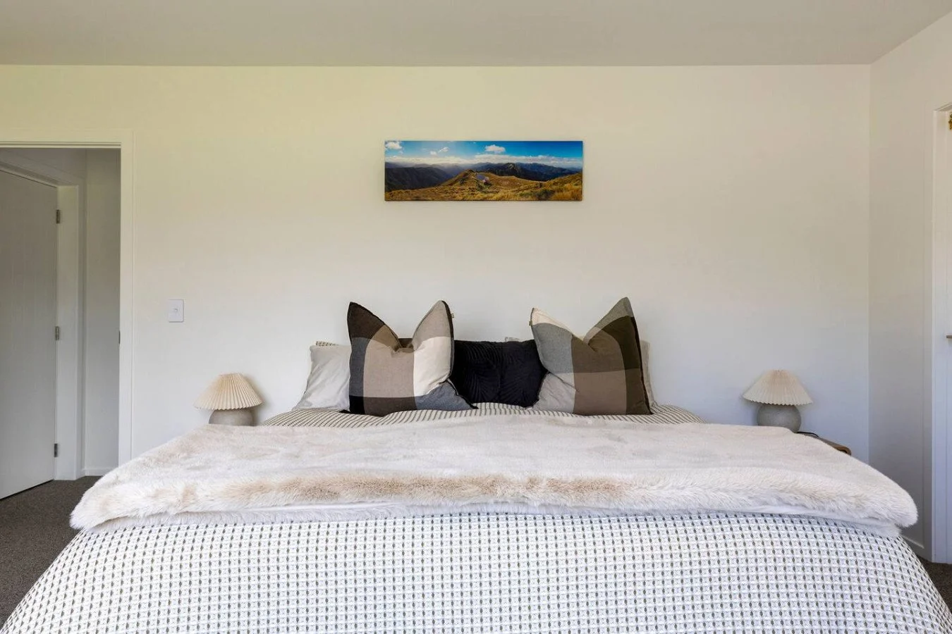 A bedroom with a large bed, dark and light checkered pillows, white lamps on bedside tables, a mountain landscape photograph on the white wall.