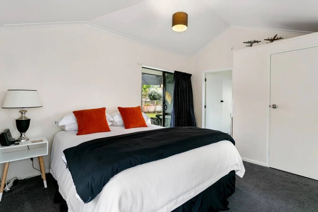 Bedroom with a bed featuring white sheets, black blanket, and two orange pillows. Side table with a lamp, phone, and remote. Sliding glass door with a view of a backyard and a black curtain. Ceiling light fixture shaped like a drum. Decorative model 