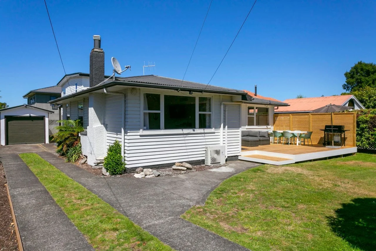 Backyard of a house with a deck, outdoor dining table, grill, and seating area, with a small lawn and driveway.