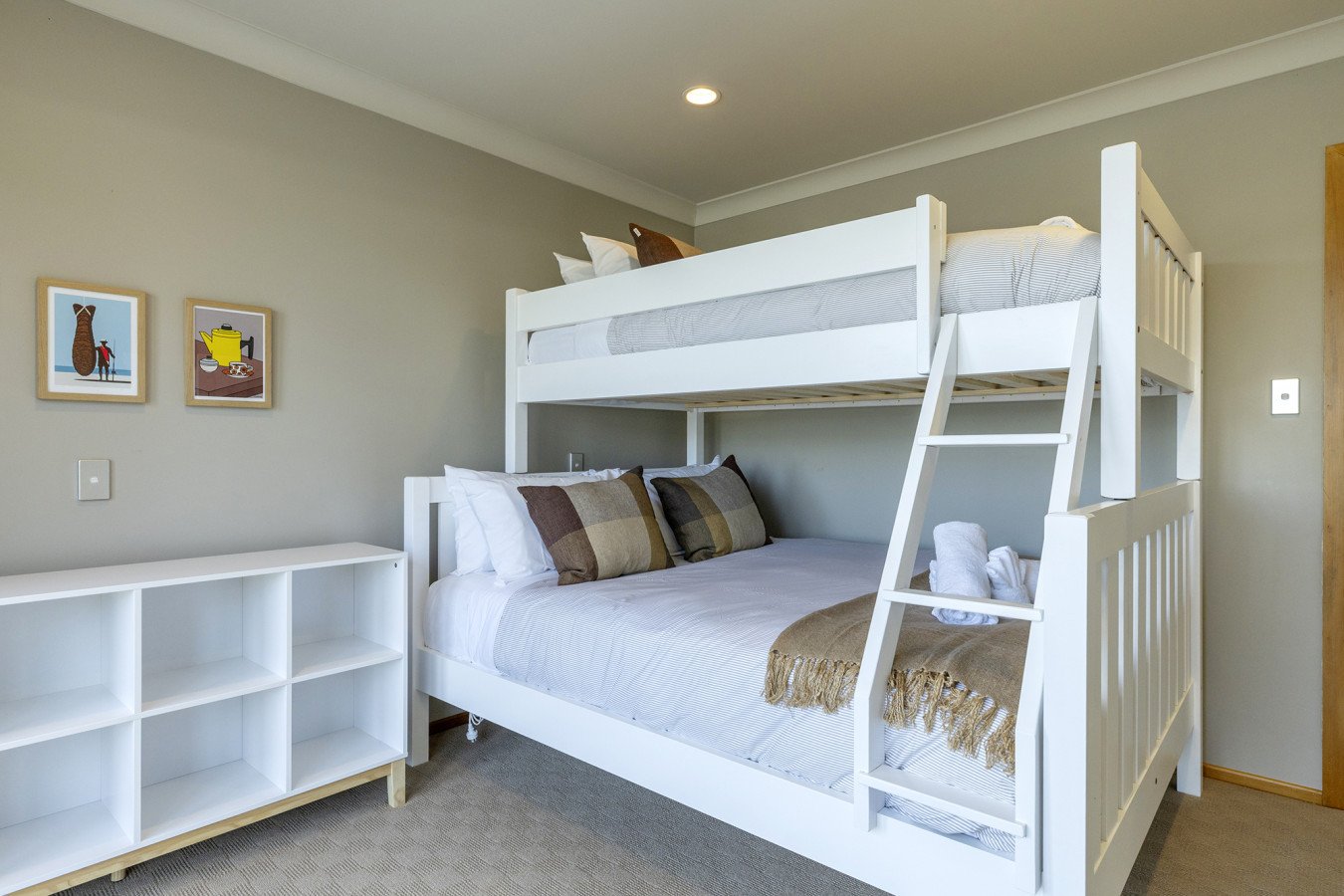 A bedroom with a white bunk bed and a double bed beneath. The double bed has white bedding, pillows, and a brown plaid blanket. The bunk bed has a ladder. On the wall, there are three framed pictures.