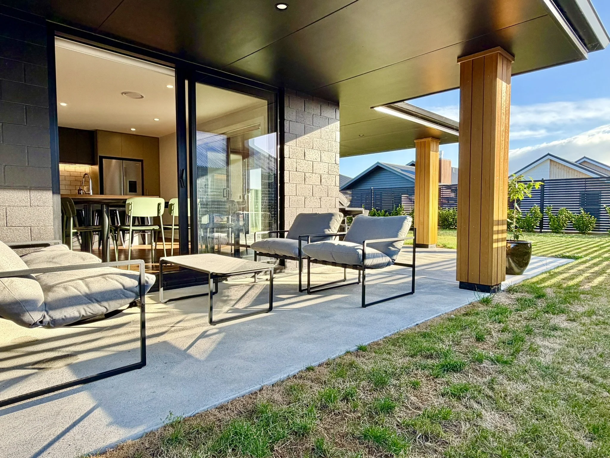 Modern outdoor patio with cushioned chairs and coffee table, overlooking a backyard with grass and houses, adjacent to a house with large sliding glass doors providing indoor access.