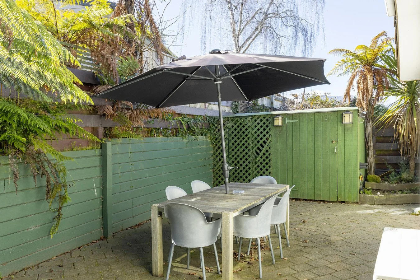 Outdoor patio with a rectangular wooden table, six gray chairs, and a large black patio umbrella. There are green wooden fences and a small green shed in the background, with some greenery and trees.