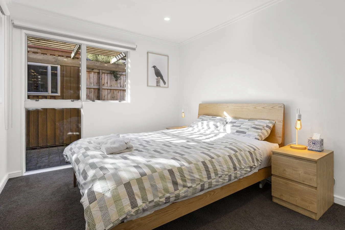 A bedroom with a wooden bed frame and a checkered bedspread. There is a nightstand with a small lamp, tissue box, and an on/off switch. A framed picture of a bird hangs on the white wall. The window shows a view of a wooden fence and outdoor space.