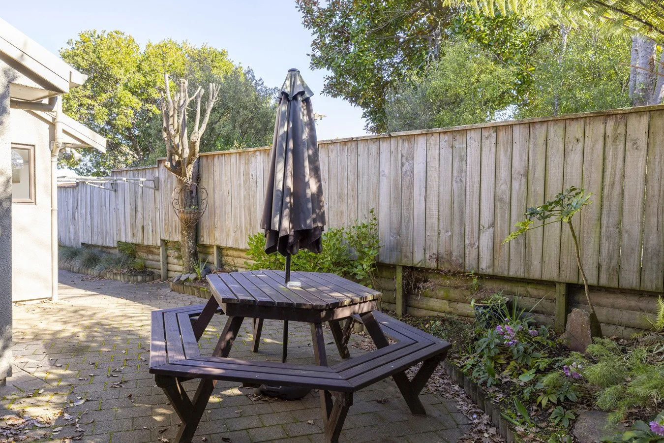 A backyard patio with a round wooden picnic table and a black umbrella, bordered by a wooden fence and garden with lush green plants and flowers.