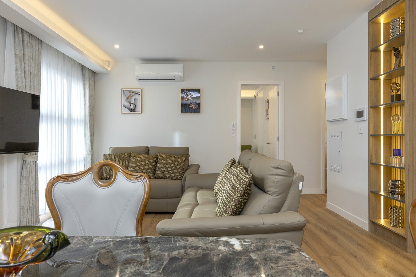 Living room with beige sofas, patterned cushions, wall art, a mounted TV, a glass bowl, a marble table, wooden flooring, and a decorative bookshelf with spotlights.
