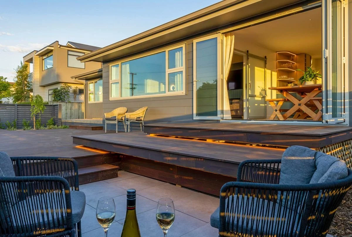 A modern house with an outdoor patio area featuring two wine glasses and a bottle of wine on a table, with chairs, a wooden deck, and large sliding glass doors leading inside the house.