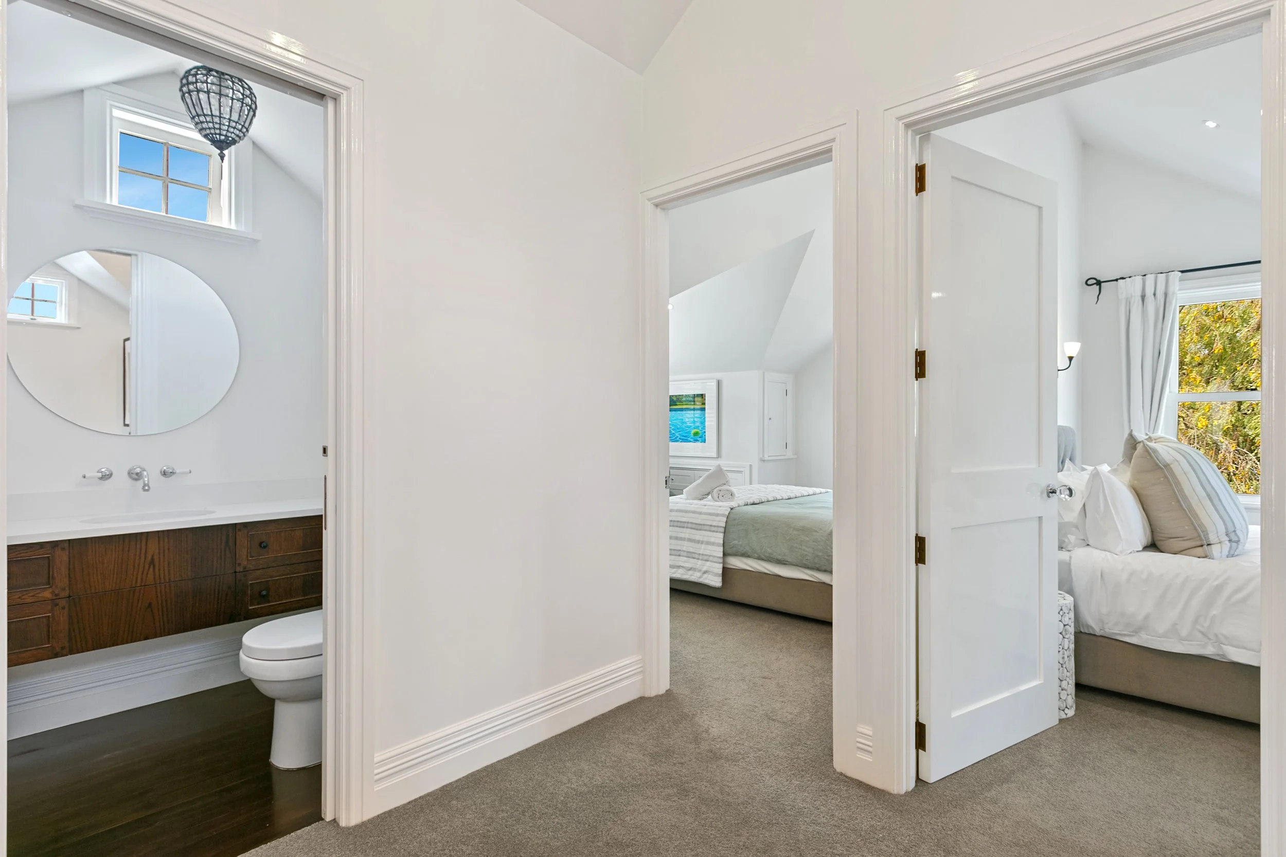 Interior view of a house showing a hallway with two bedrooms visible through open doors and a bathroom on the left. The bedrooms have beds with pillows and windows with curtains, and there is a decorative light fixture above the bathroom.