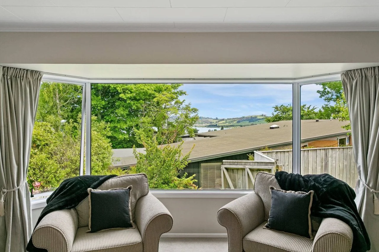 Living room with large window overlooking green trees, rooftops, and hills in the distance. Two beige striped armchairs with black pillows and black throws are positioned in front of the window, with cream curtains on either side.