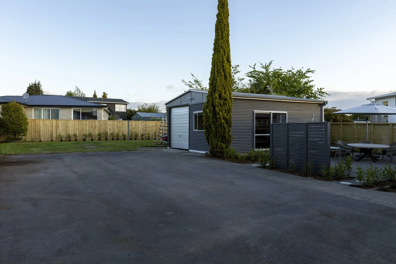 View of a residential backyard with a small gray shed, tall tree, patio furniture with an umbrella, and a wooden fence, during daytime.