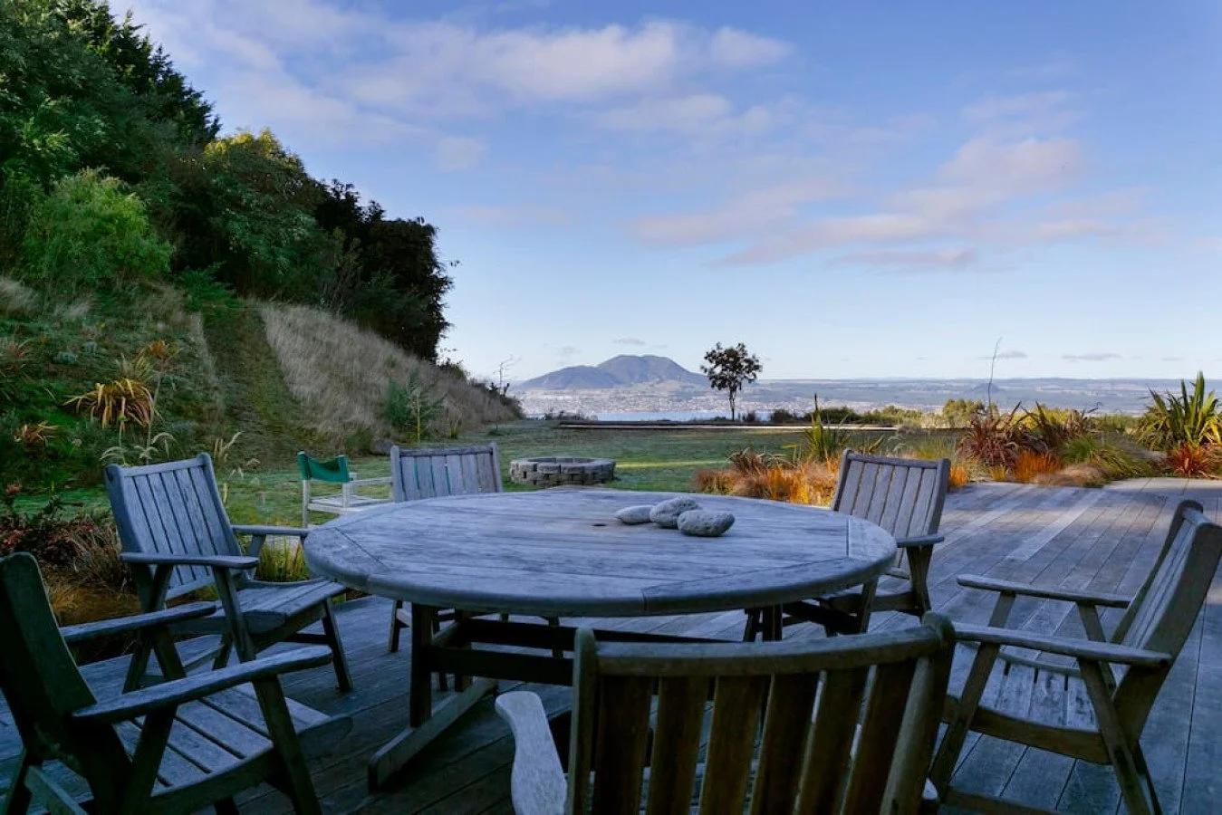 Outdoor patio with a round wooden table and six chairs, surrounded by landscaping, trees, and a distant view of a lake and mountains under a partly cloudy sky.