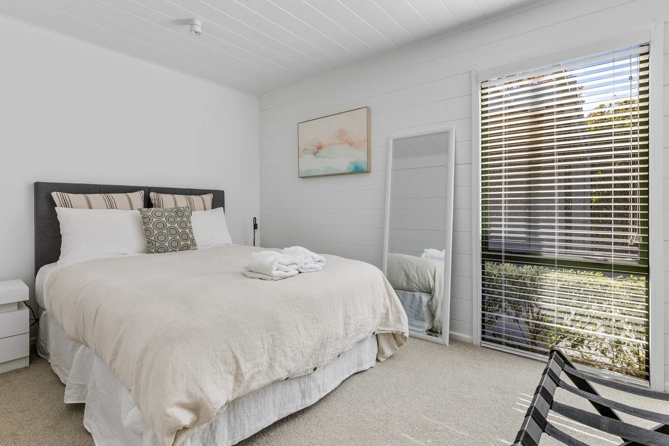 A cozy bedroom with a large bed, white linens, and decorative pillows, next to a window with blinds and a full-length mirror leaning against the wall.