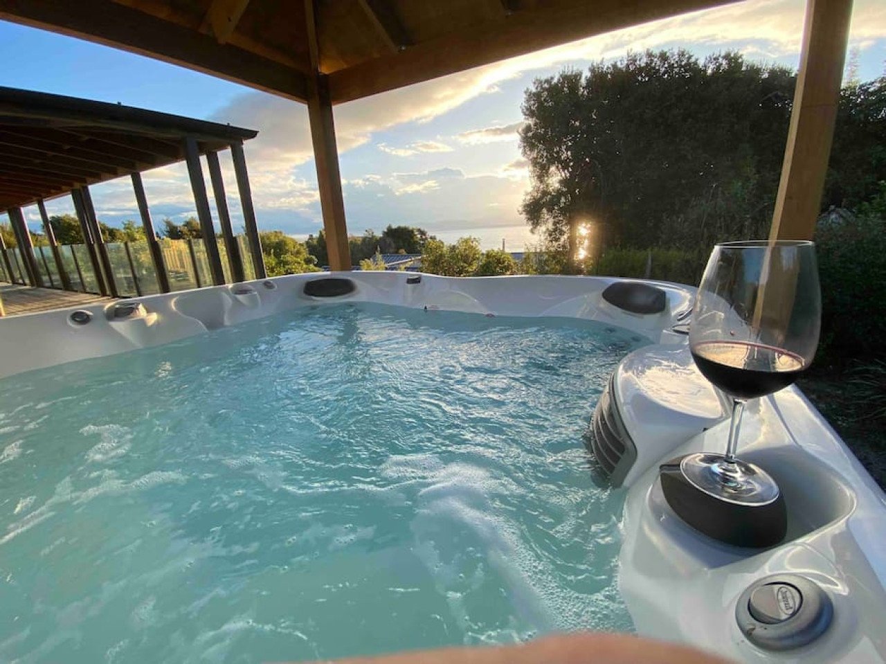 A hot tub filled with water, with a glass of red wine resting on the edge, under a wooden roof with scenic view of trees, ocean, and sunset in the distance.