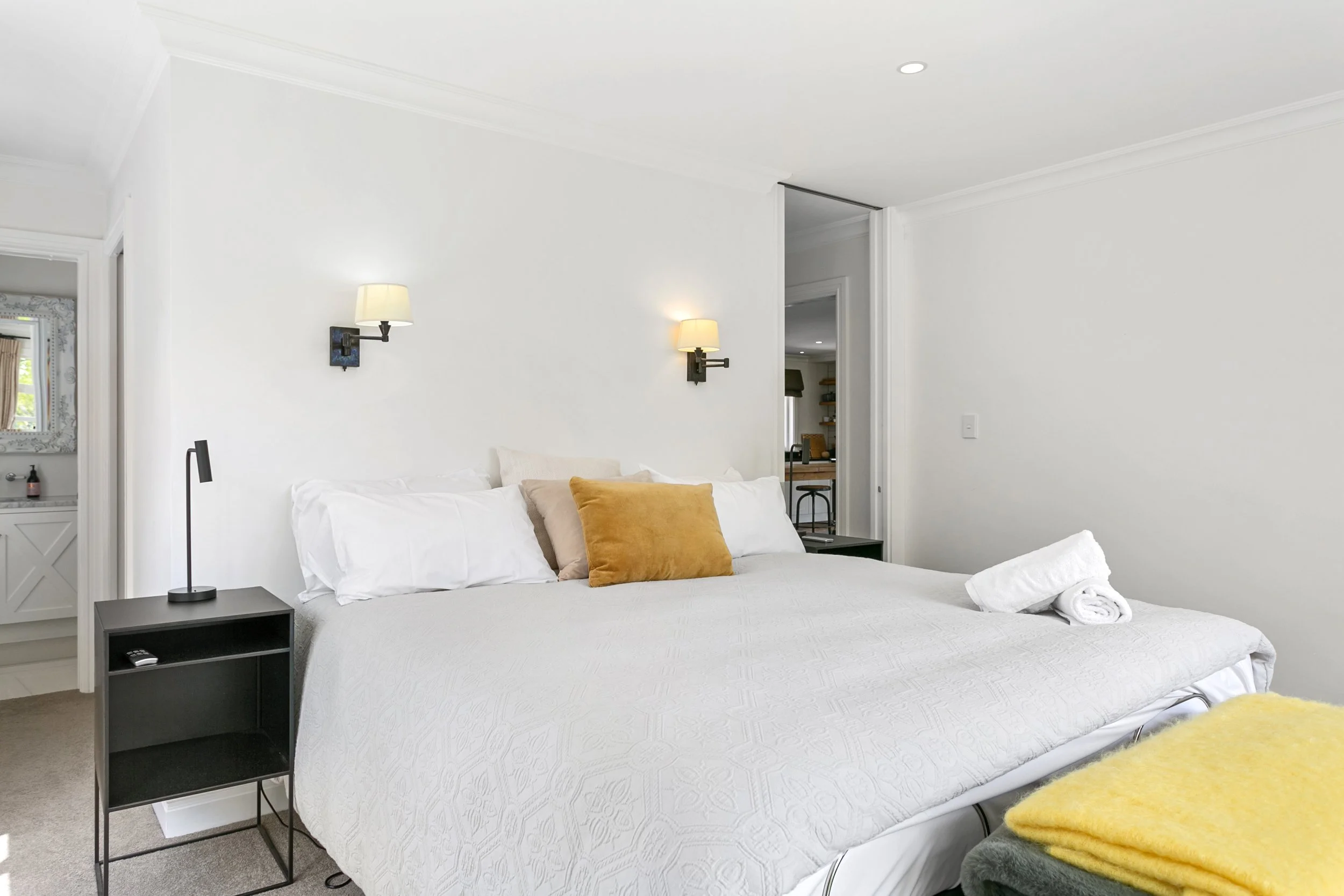 A neatly made bed with white bedding, a mustard yellow pillow, and white towels rolled on the side. Two wall-mounted lamps above the bed, with a small black nightstand on each side. A glimpse of a bathroom with a mirror and vanity is visible on the l