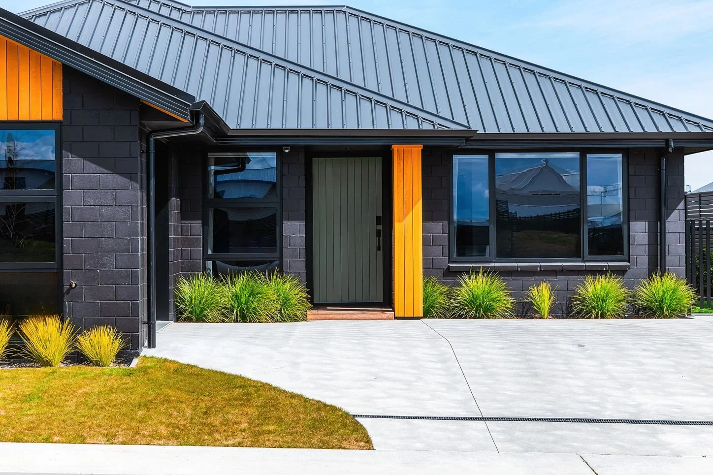 Front view of a modern house with black brick walls, large windows, a black metal roof, and a wooden accent on the doorframe. There are plants along the sidewalk and a lawn in the foreground.