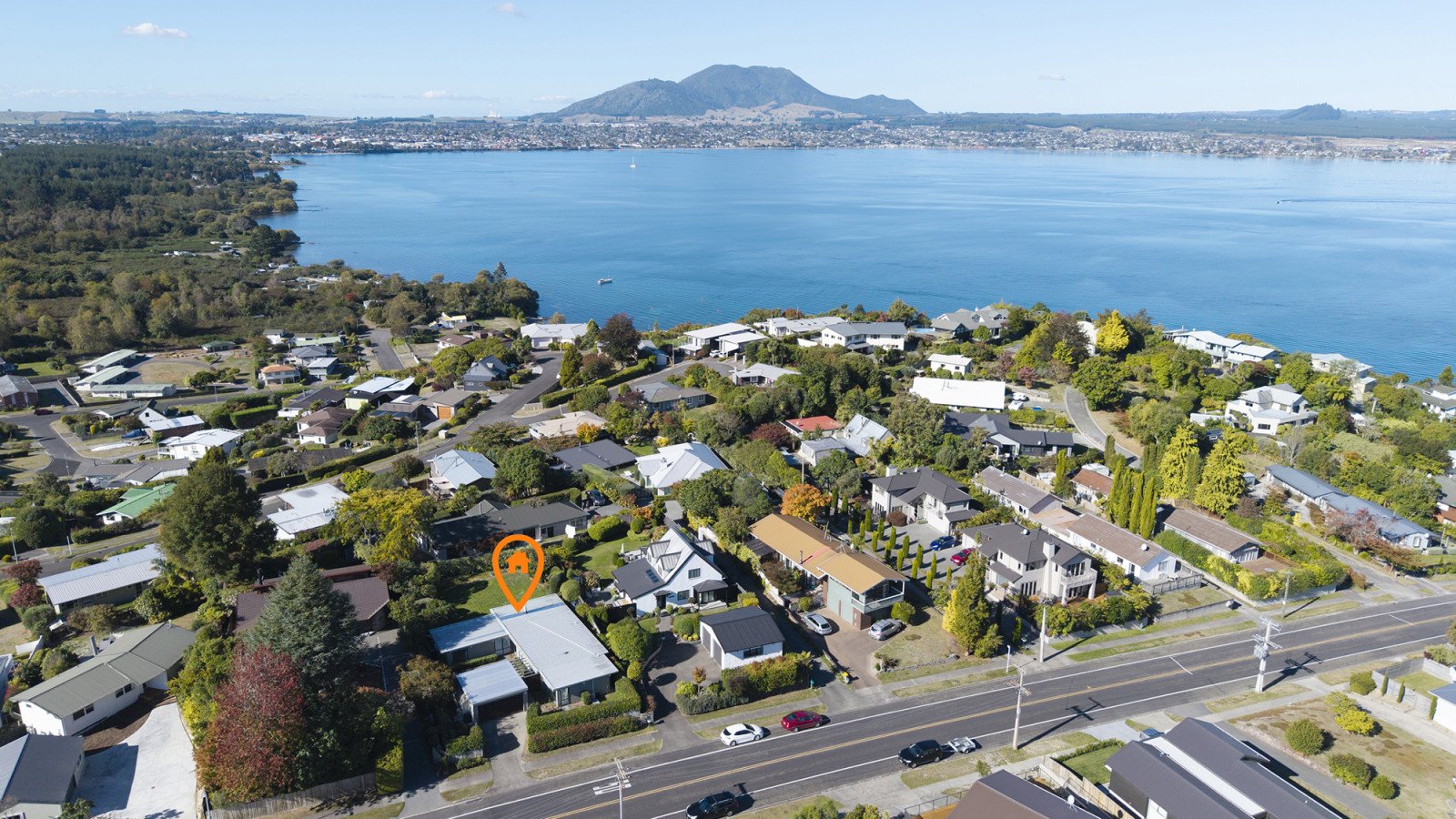 Aerial view of a residential neighborhood near a large body of water with mountains in the background. The neighborhood has houses, trees, and roads.