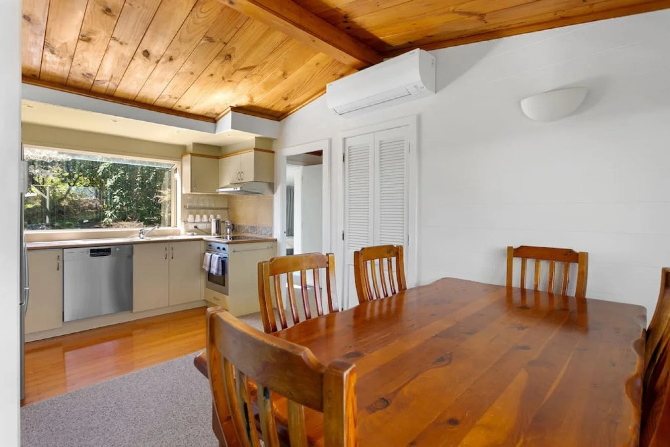 Dining room with a large wooden table and six matching chairs, adjacent to a kitchen with beige cabinets, a large window, and wooden ceiling panels.
