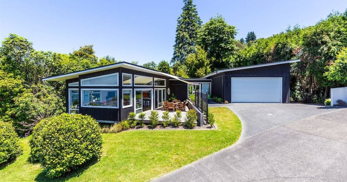 Modern house with large glass windows, black exterior, surrounded by green trees, with a curved driveway and a small outdoor patio with a table and chairs.