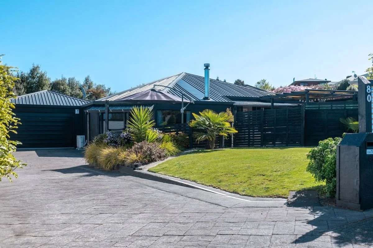 A modern house with a metal roof, surrounded by a well-maintained lawn, palm trees, and shrubs, with a paved driveway leading to the garage.