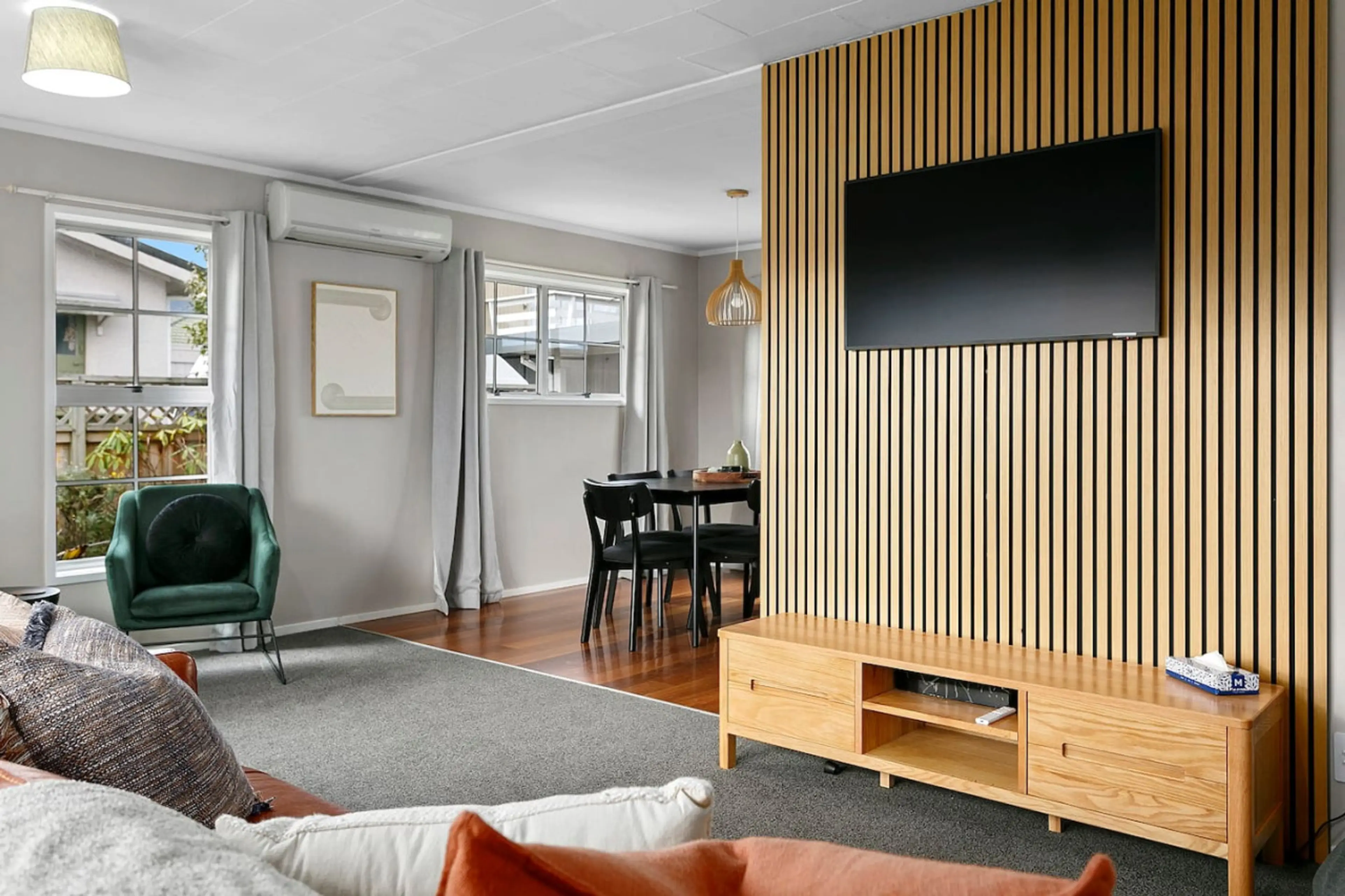 Living room with a TV mounted on a wooden slat wall, a green armchair near the window, a dining table with black chairs, and a wooden console table.