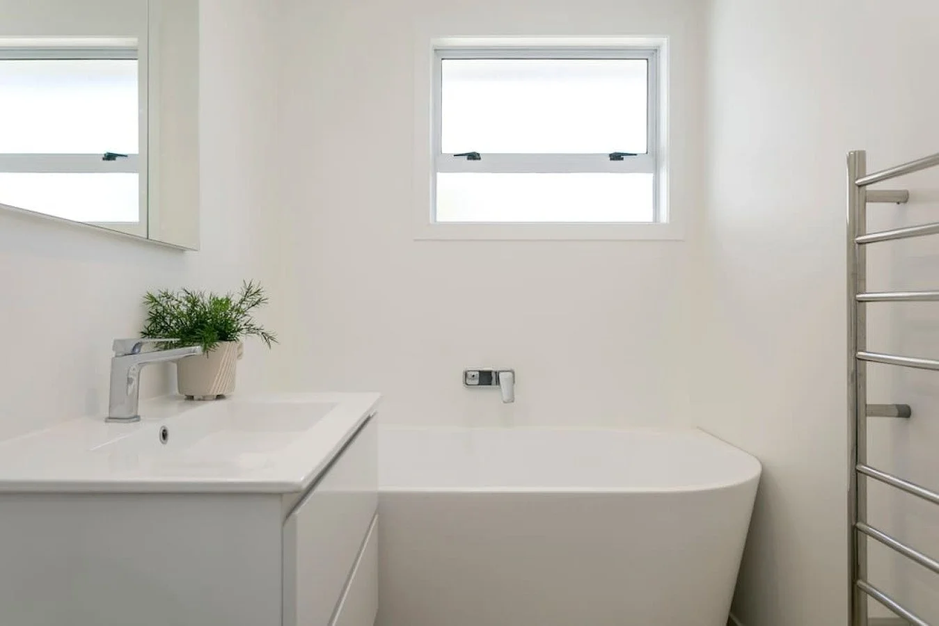 A minimalist bathroom with a white bathtub, a white vanity with a plant, a window, and a heating towel rack.