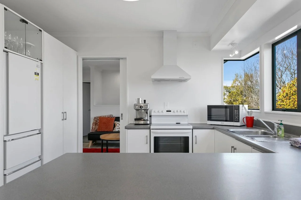 Modern kitchen with white cabinets, gray countertops, a microwave, coffee maker, stove, and a view of trees through large windows.