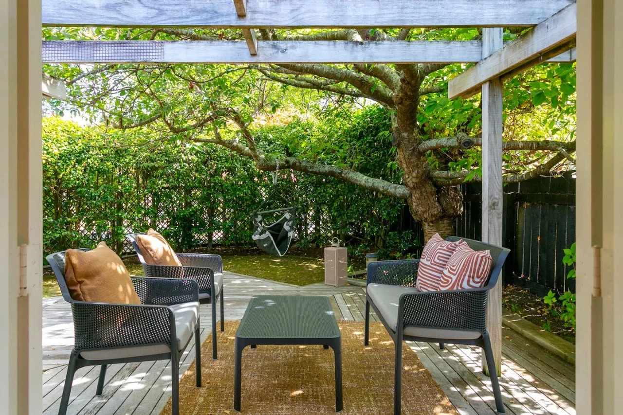 Patio with outdoor furniture, a small table, four chairs with cushions, a swing hanging from a tree, trees, bushes, and a black fence.