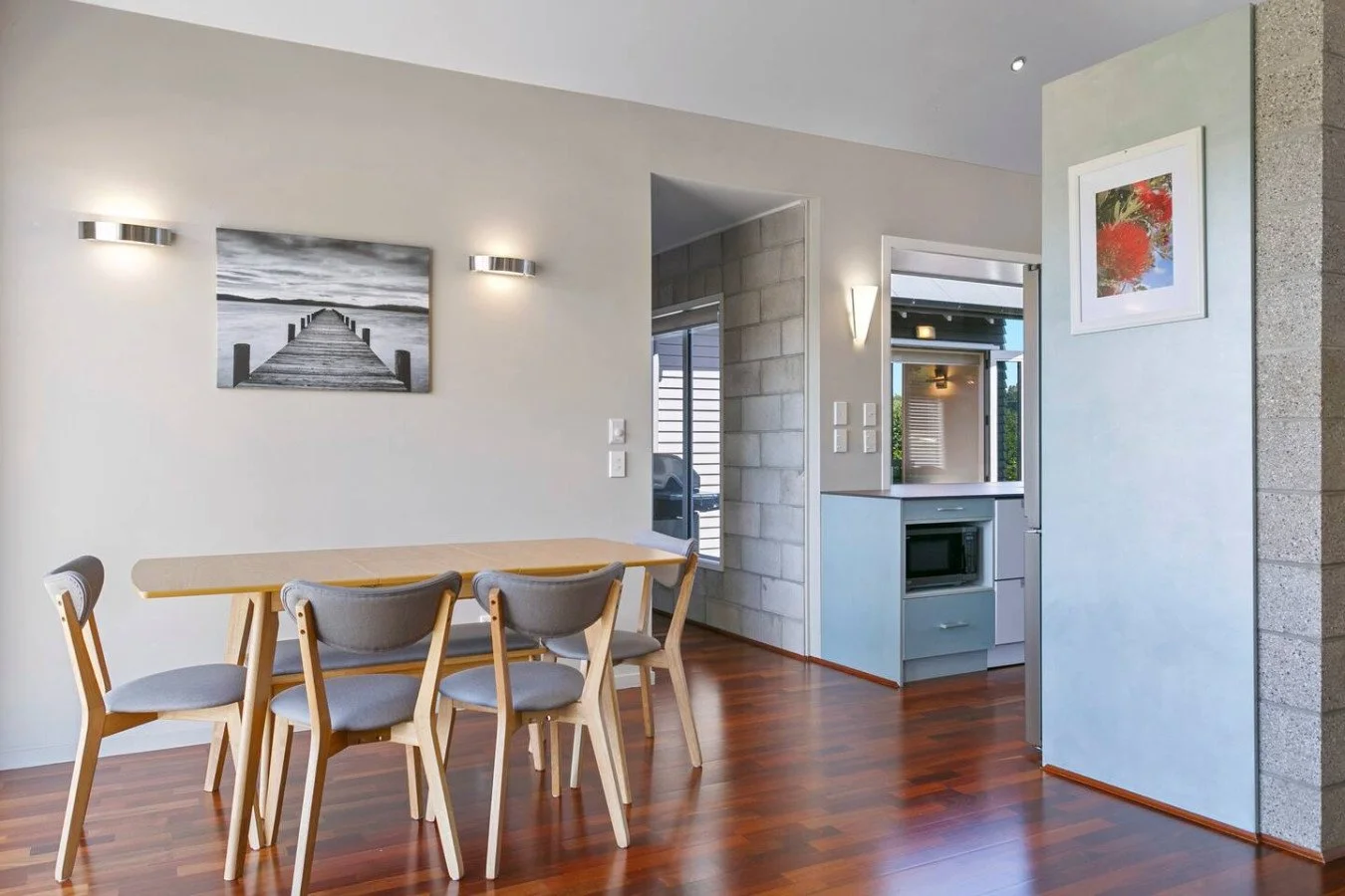 Dining area with a wooden table and six gray chairs, artwork on the wall, hardwood flooring, and a view into the kitchen with a microwave and a small window.