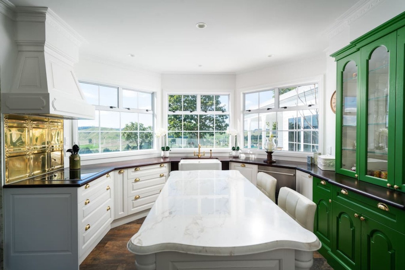 Bright kitchen with white cabinets, dark countertops, and green cabinet. Large windows overlooking a scenic landscape, central white island, and farmhouse sink.
