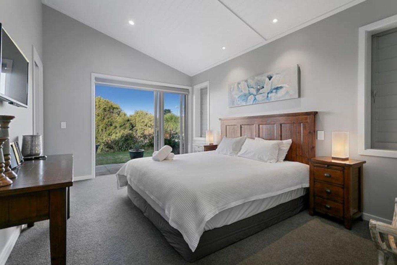 Modern bedroom with a large bed, wooden headboard, nightstands with lamps, artwork above the bed, sliding glass door leading to a garden, and a TV on a wooden dresser.