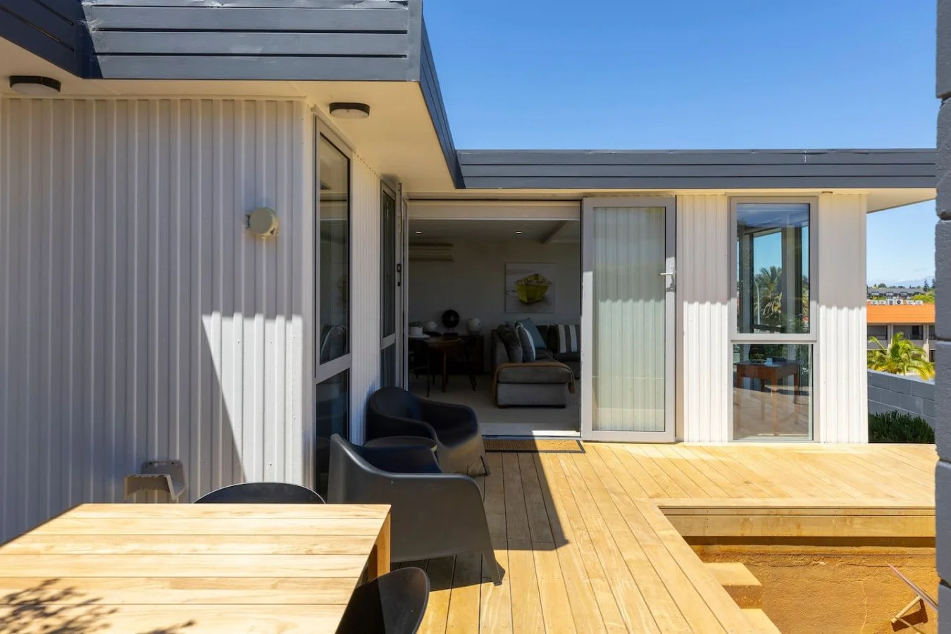 Exterior view of a modern house with a wooden deck, partially open sliding glass door leading to a living room, and outdoor furniture including black chairs and a wooden table under a blue sky.