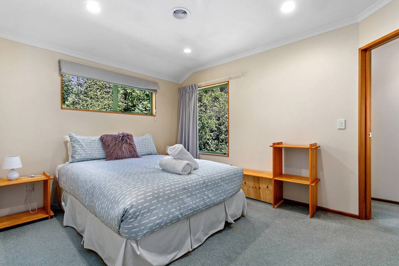 Bedroom with bed, two rolled towels, and decorative pillows, two windows overlooking greenery, side table with lamp, small wooden shelf, light-colored walls, and carpeted floor.