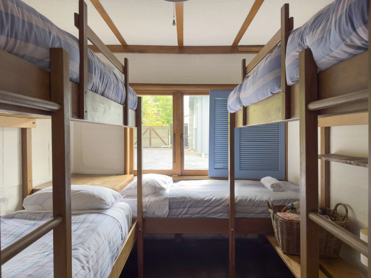 A room with wooden bunk beds, blue shutters on the door, and a window with a view of outside greenery.