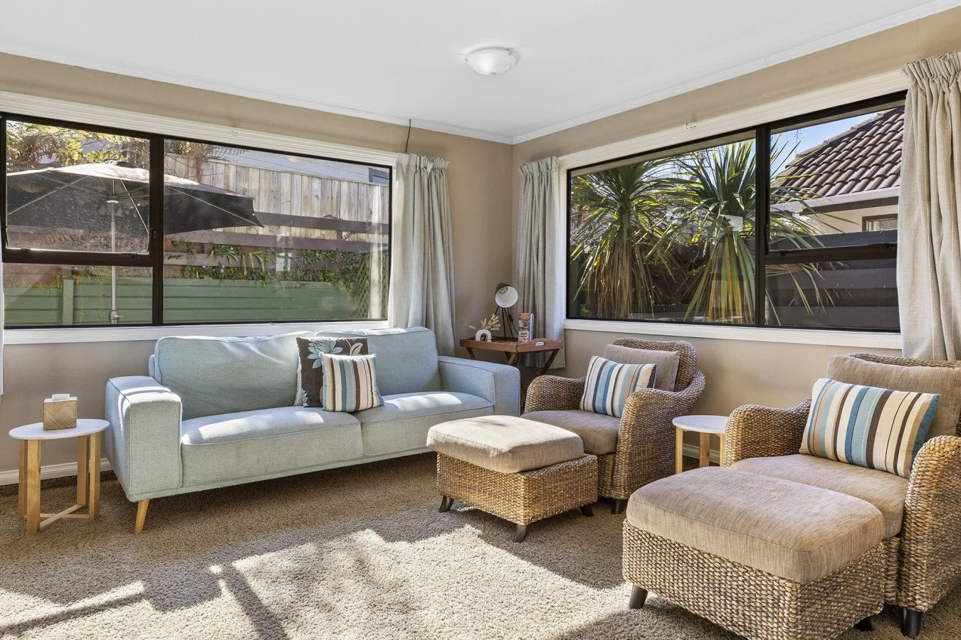 Living room with beige walls, large windows with cream curtains, light blue sofa, wicker armchairs with striped pillows, small side tables, a brown desk with a white lamp and decor, and a beige carpet.