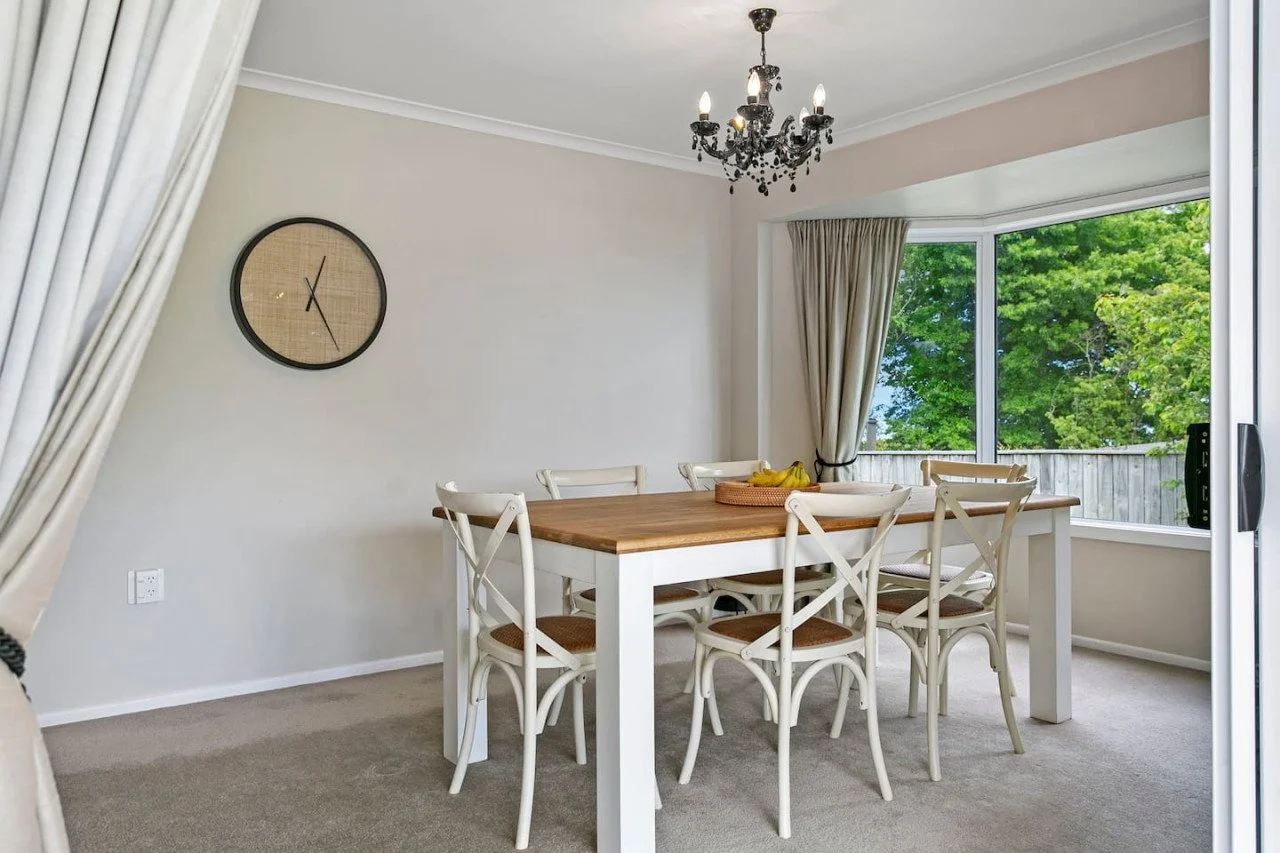 Dining room with a wooden table, six white chairs, a black chandelier, a large wall clock, beige curtains, and a window showing green trees outside.