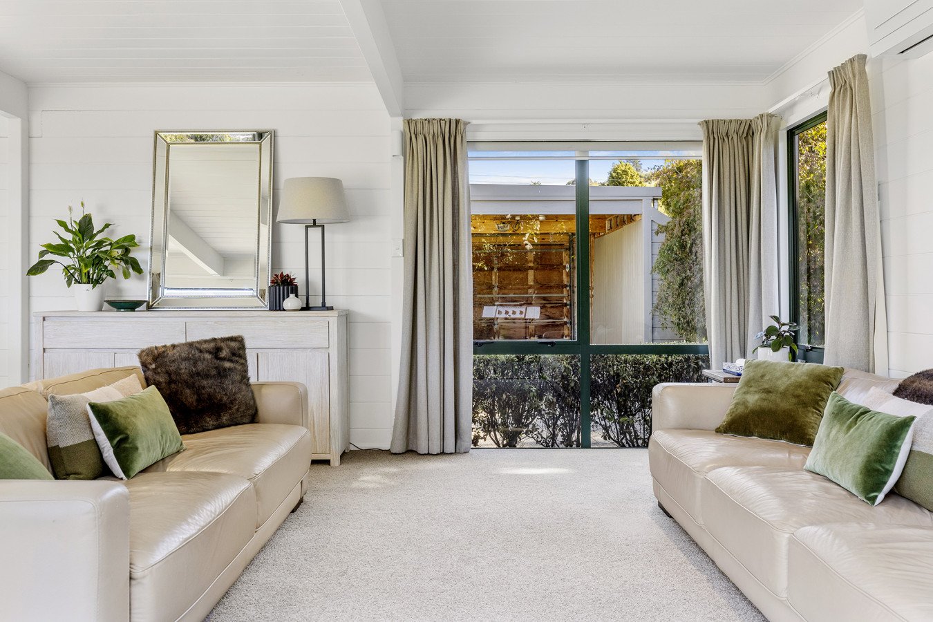 Living room with cream leather sofas, green and brown pillows, a white sideboard, a large mirror, potted plants, a window with curtains showing a backyard, and a carpeted floor.