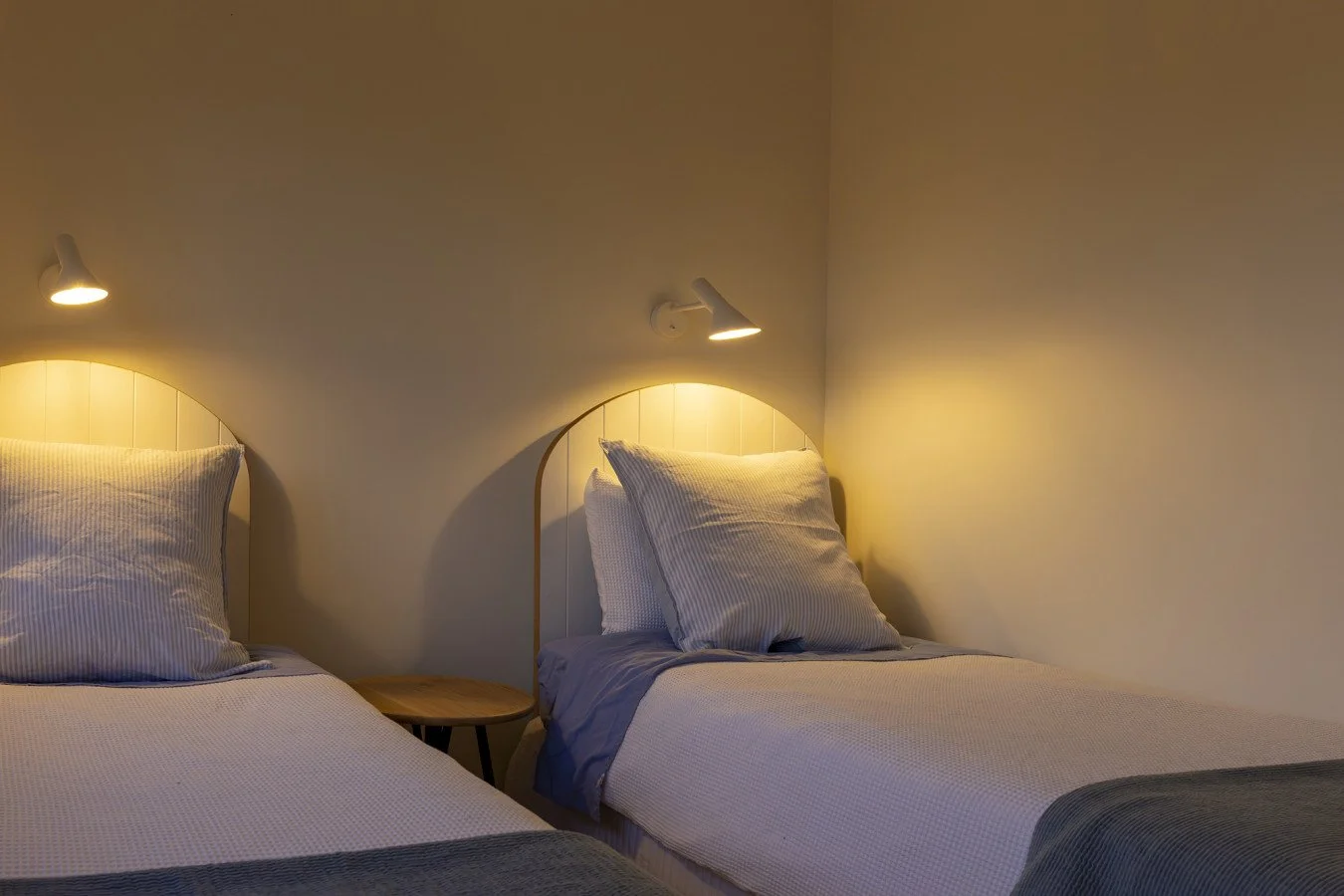 Two single beds with striped pillows and linens, a small round wooden bedside table between, and wall-mounted reading lights in a cozy bedroom.