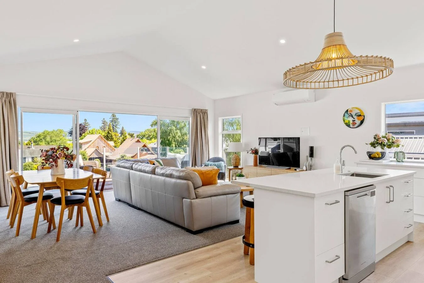 Open-plan living and dining area with large windows showing a neighborhood view, white walls, beige carpet, a white leather sofa, a wooden dining table with chairs, a television, and a kitchen island with a sink and flowers.