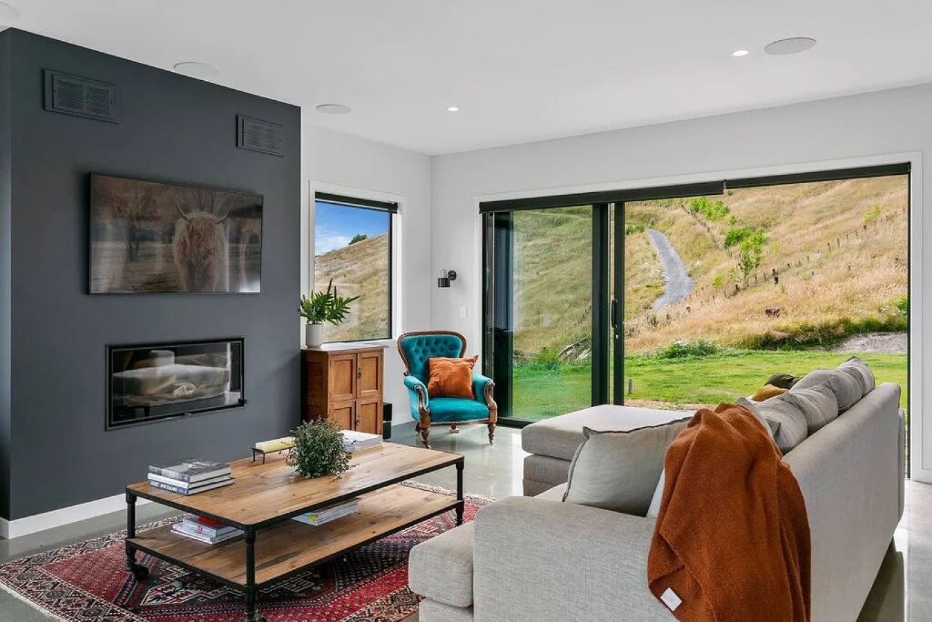 Living room with large sliding glass doors showing a hillside landscape, a beige sectional sofa with orange throw blanket, a wooden coffee table with books, a blue armchair, and a TV mounted on a dark gray accent wall with a fireplace underneath.