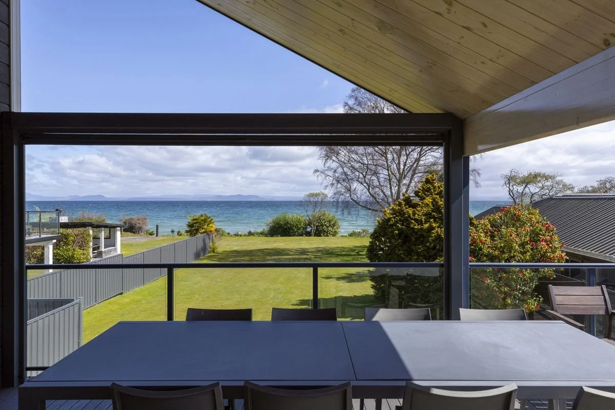 View from a covered porch looking out over a grassy yard towards the ocean, with trees and shrubs, and a cloudy sky in the distance.