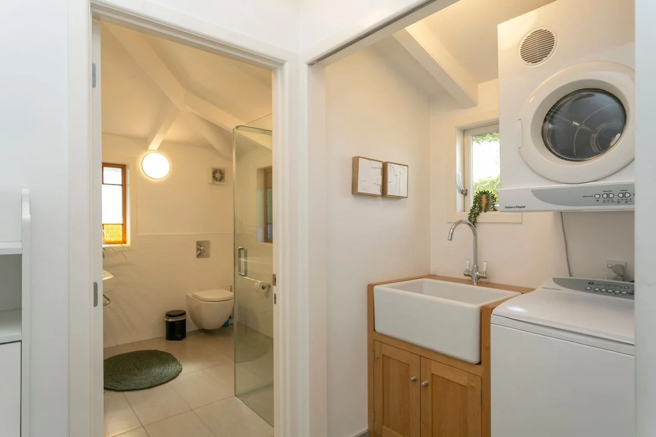 A combined laundry and utility room with a washer and dryer stacked, a small window, a white utility sink with wooden cabinet, and a wall-mounted bathroom visible through an open door.
