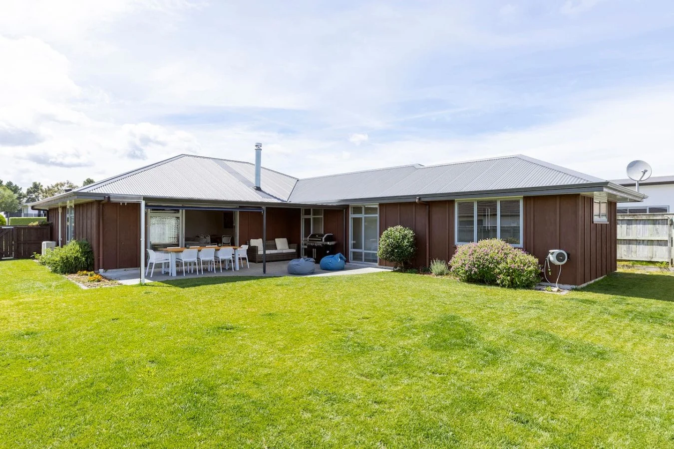 Backyard of a brown house with an outdoor dining area, lounge chairs, grill, and lush green lawn under a partly cloudy sky.
