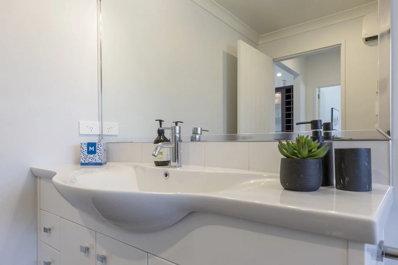 Bathroom sink with black soap dispenser, plant in black pot, and black cup on a white countertop; large mirror and white walls.