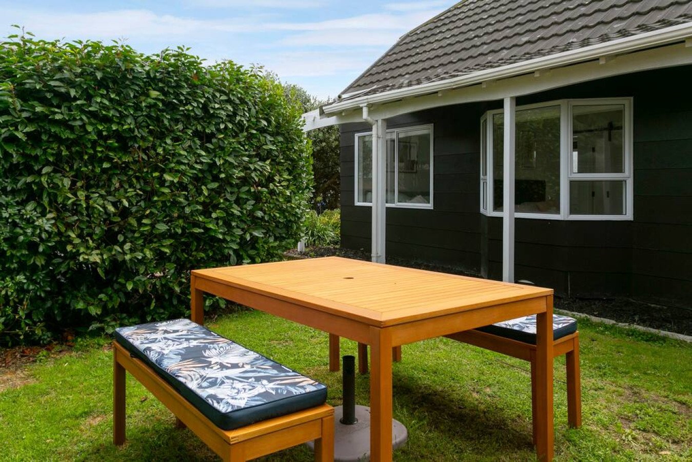 A patio table with two benches, one with a floral cushion, on a green lawn next to a black house with white framed windows and a hedge.
